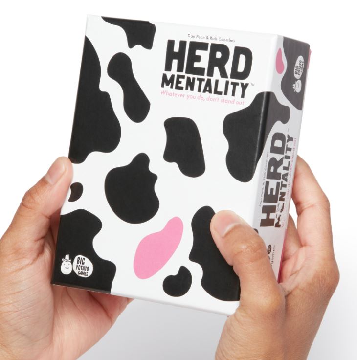 Two hands are holding a box designed with a cow print pattern in black, white, and pink. The text on the box reads, "HERD MENTALITY MINI" and "Whatever you do, don't stand out! BIG family game." Perfect for gatherings, this party game from Blue Dog Posters features playful cow graphics and branding.