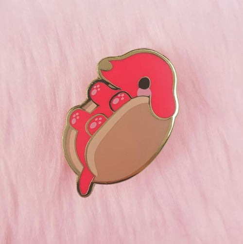 Introducing the Hot Dog Pin Pinstachiio by Blue Dog Posters: a delightful enamel pin showcasing an adorable red animal character with a curled tail and tiny paws, nestled in a cozy brown shell-like structure. Set against a soft pink, furry background, this charming accessory is perfect for adding flair to your Ita bag or collection in Wollongong, Australia.