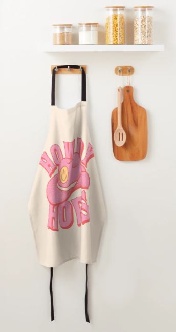 A kitchen wall showcases the Howdy Hoes Cowboy Apron by Blue Dog Posters, adorned with a cartoon pink cowboy hat and the text "Howdy How's," hanging from a wooden hook. Above it, a white shelf holds three glass jars filled with pasta and grains. Next to the apron, a wooden cutting board and spoon are mounted on the wall.