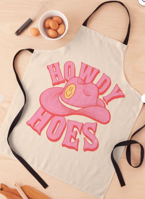 The Howdy Hoes Cowboy Apron from Blue Dog Posters' Homewares Collection, featuring black straps, is displayed on a wooden surface. Crafted from durable polyester, it showcases a playful design with a pink cowboy hat, a yellow smiley face, and the phrase "Howdy Hoes" in bold, pink Western-style text. Nearby are eggs, a whisk, and a spatula.