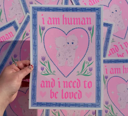 A hand holds an "i am human A4 Risograph - Amy Hastings" poster by Blue Dog Posters, featuring a pink heart with cartoon animals hugging inside. The text reads "I am human and I need to be loved" in red and pink, with a border of purple tulips and a blue geometric pattern. Other posters are scattered below.