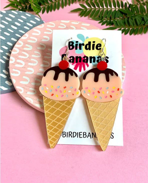 Displayed on a Blue Dog Posters card, the Ice Cream Acrylic Earrings feature a pair of handmade, colorful ice cream cones adorned with pink ice cream, chocolate sauce, a cherry on top, and sprinkles. The background showcases a pink surface accompanied by a plant and patterned plates.