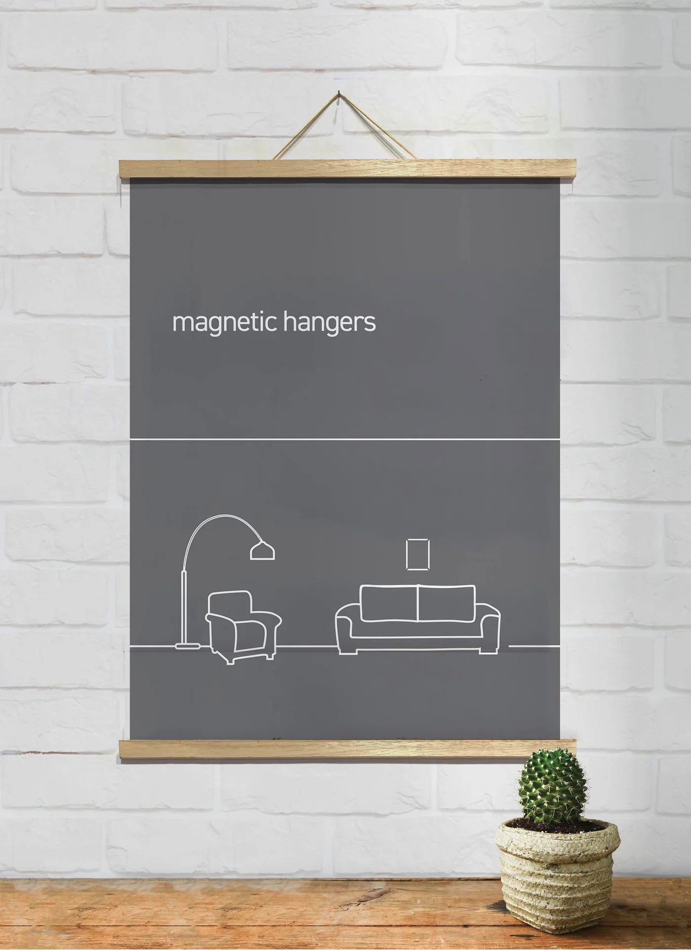 A minimalist poster hangs on a white brick wall, featuring a graphic of a living room setup with a couch, floor lamp, and picture frame. The text "Magnetic Timber Hanger 51cm Natural Pair by Blue Dog Posters" is displayed at the top. Below, a small potted cactus sits on the floor.