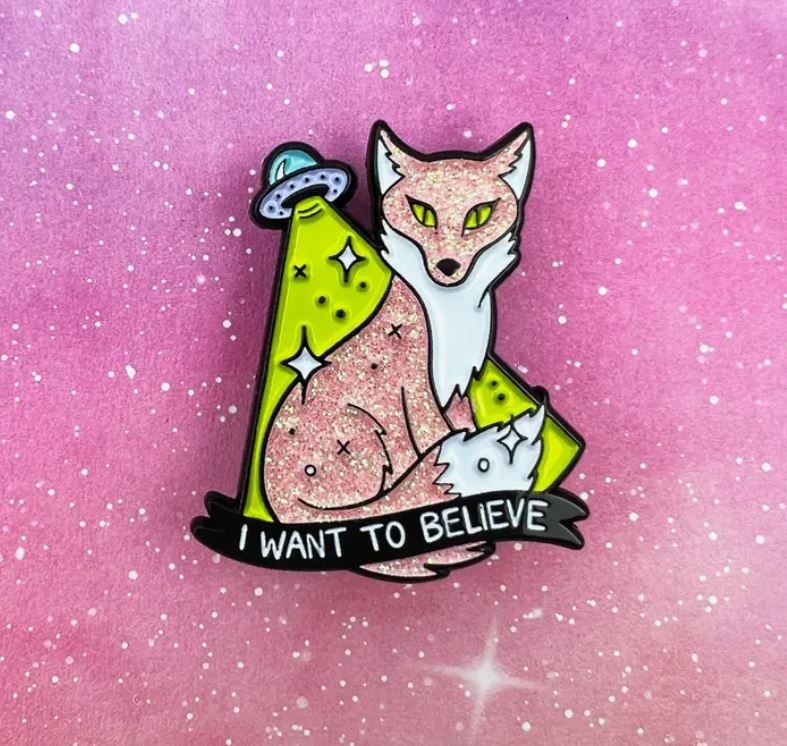 Introducing the "I Want to Believe Fox Enamel Pin," a die struck metal enamel pin featuring a sparkly pink fox with alien-like eyes sitting in front of a cartoon-style UFO that emits green light. Crafted by Blue Dog Posters, this charming accessory showcases the text "I WANT TO BELIEVE" on a ribbon below the fox. The background boasts a captivating pink and purple gradient adorned with small white stars.