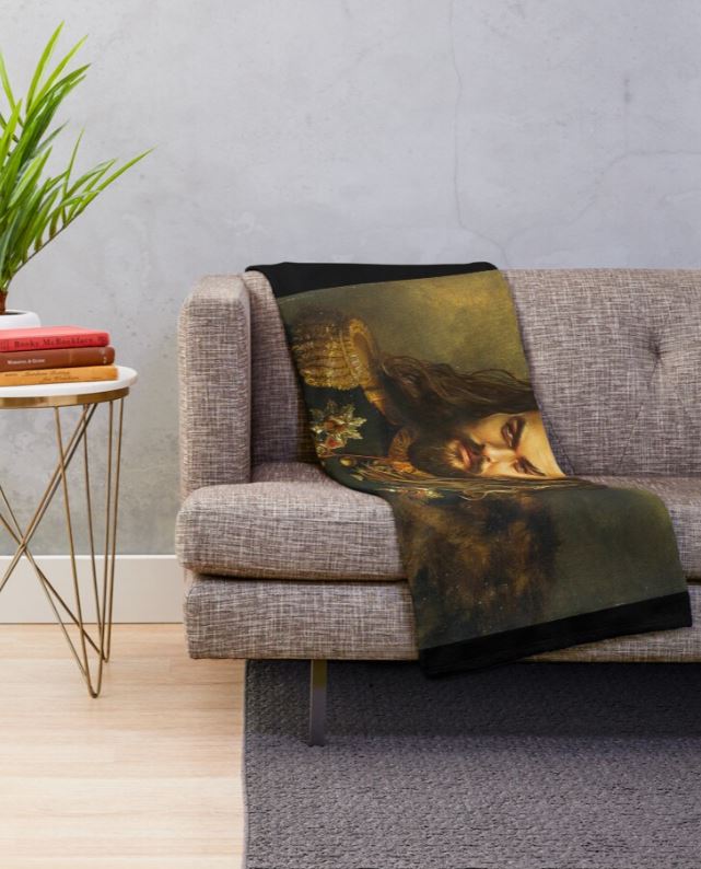 A gray couch has a soft, fluffy "Jason Momoa Replace Face Throw Blanket" by Blue Dog Posters draped over the armrest, featuring a detailed painting of a bearded man with long hair. The couch is next to a small round side table with a stack of books and a potted plant. The scene is set against a simple wall with a gray texture.