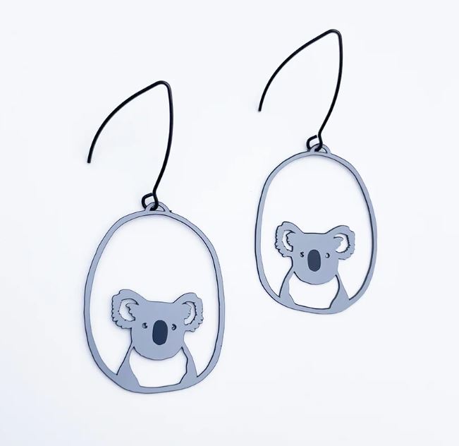 The Denz Mini Earrings Koalas Grey, designed by Blue Dog Posters, elegantly feature cut-out koala face designs framed in an oval shape. Suspended from surgical steel hooks and crafted from recycled plated stainless steel, these adorable earrings celebrate Australia against a plain white background.