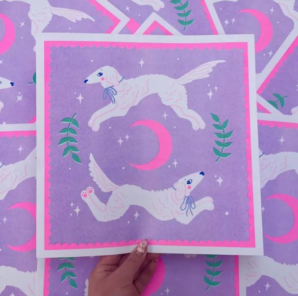 The "Leaping Borzois Risograph" by Amy Hastings from Blue Dog Posters features two white dogs with blue bows jumping around a pink crescent moon, set against a purple background with stars and green leaves. A hand holds the square artwork, matching the patterned paper beneath it.
