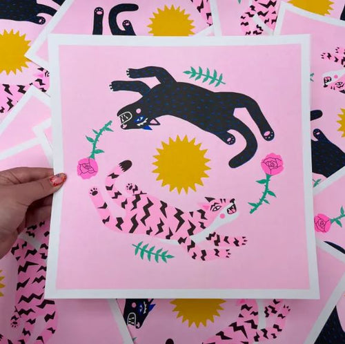 A person holds the Leaping Cats Risograph by Amy Hastings from Blue Dog Posters, featuring a vibrant design with a black panther and pink zigzag-striped panther circling a yellow sun amidst pink roses and green leaves on a pink background.