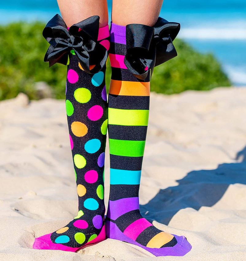 On a sandy beach, someone strikes a pose showing off their Madmia Liquorice Bows Socks from Blue Dog Posters. One sock is decorated with vibrant dots, and the other displays bold stripes, both topped with oversized black bows. The tranquil ocean and lush greenery form a serene backdrop to this playful scene.