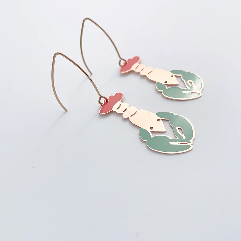 Introducing the Lobster in Coral Denz Earrings by Blue Dog Posters: These unique earrings are shaped like hands forming different gestures, adorned with vibrant red and green enamel accented by elegant gold outlines, echoing the shape of lobster claws. Designed with a minimalist hook for comfortable wear, these lightweight statement pieces are perfect against a light gray background.