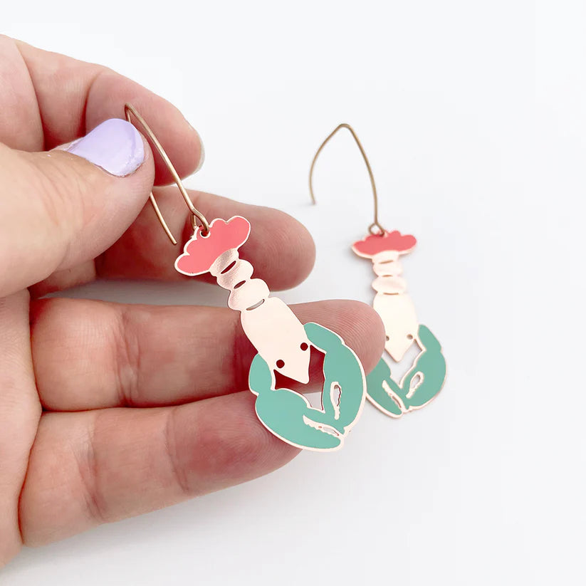 A hand holds a Lobster in Coral Denz Earring featuring pink and turquoise enamel with a gold hook—an exquisite piece from Blue Dog Posters. Another lightweight midi lobster earring lies elegantly in the background on a white surface.