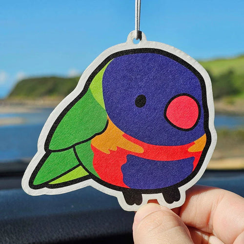 A hand holds up the Blue Dog Posters Lorikeet Bubble Gum Air Freshener, a colorful parrot-shaped accessory with purple, green, red, and blue sections, against a blurred outdoor backdrop seen through a car window.