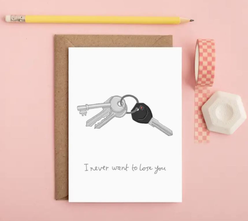 The Never Want to Lose You Greeting Card by Blue Dog Posters features a whimsical design of a set of keys on a keyring, including a car key. The card comes with a brown envelope, accompanied by a yellow pencil, checkered tape, and a white hexagonal item. It reads, "I never want to lose you." This charming greeting card adds a touch of humor to any occasion.