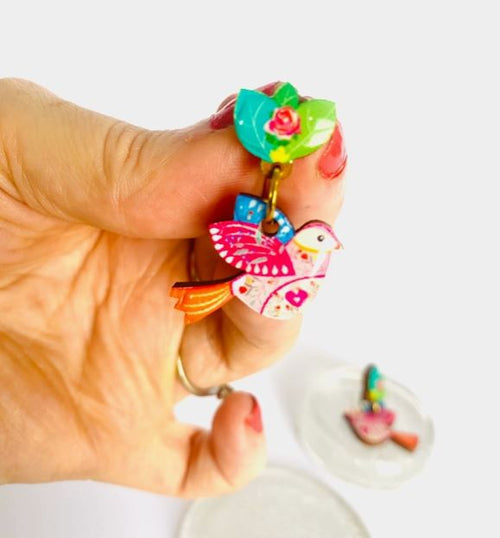 A hand with red-painted nails holds a Love Bird Retro Earring by Blue Dog Posters, featuring a colorful pink bird with patterns, an orange tail, and a green leafy top. A similar earring appears blurred in the background.