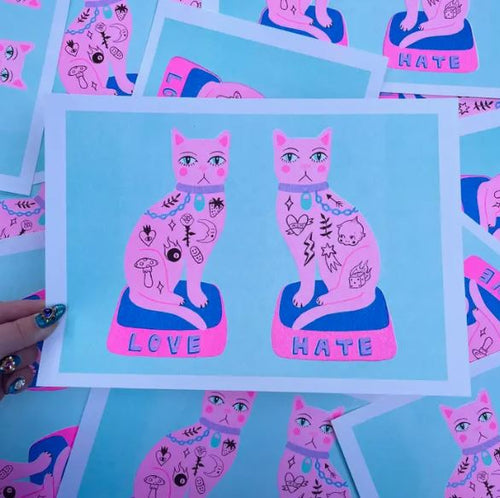 A hand displays an A4 risograph print, "Love Hate Cats" by Amy Hastings from Blue Dog Posters, depicting two tattooed pink cats on blue with bases labeled "LOVE" and "HATE." Identical prints are scattered below.
