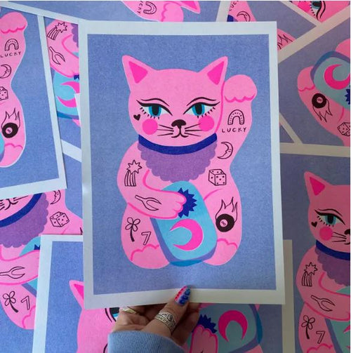 A person is holding an A4 risograph print titled "Lucky Cat" by Amy Hastings from Blue Dog Posters, which features a pink Maneki-neko cat adorned with symbols like a horseshoe, clover, star, and dice against a vibrant lavender background. Several similar prints are scattered around.