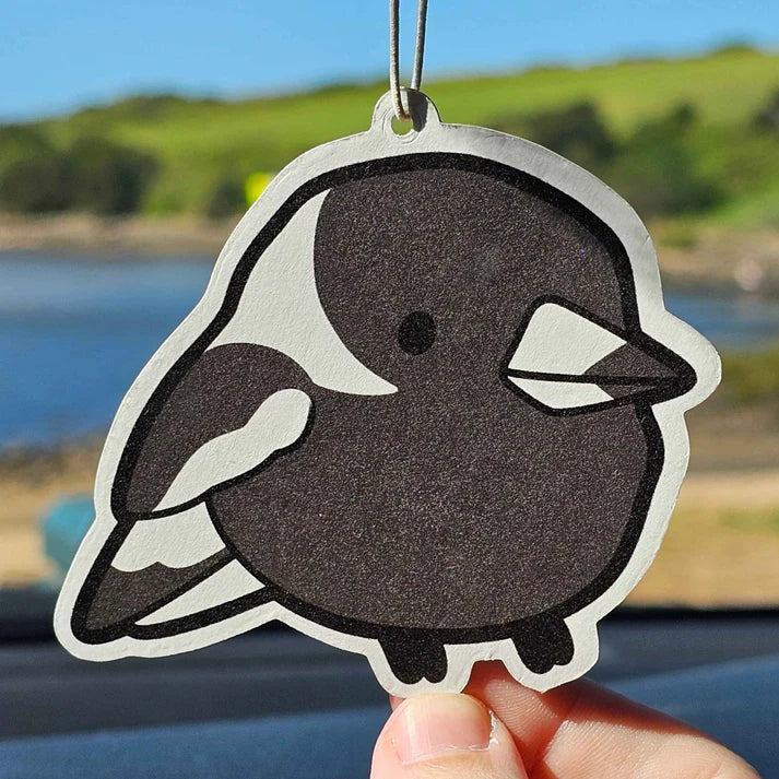 A hand holds up the Magpie Grape Air Freshener by Blue Dog Posters, a cute cartoon-style black and white bird, with a blurred outdoor background of water, grass, and trees.