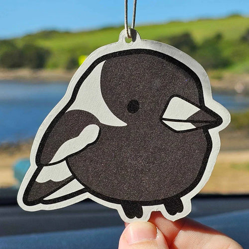A hand holds up the Magpie Grape Air Freshener by Blue Dog Posters, a cute cartoon-style black and white bird, with a blurred outdoor background of water, grass, and trees.