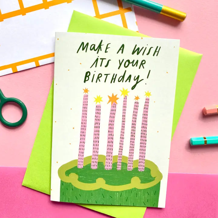 The Make a Wish Cake Greeting Card by Blue Dog Posters features a green cake with seven pink candles and the message "MAKE A WISH IT'S YOUR BIRTHDAY!" It comes with a green envelope, perfect for birthdays.