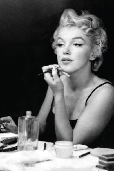 A woman with short, curly blonde hair in a black dress applies Marilyn Monroe Makeup by MTV 070 at her vanity, surrounded by makeup and bottles, creating a dramatic scene against a dark background.