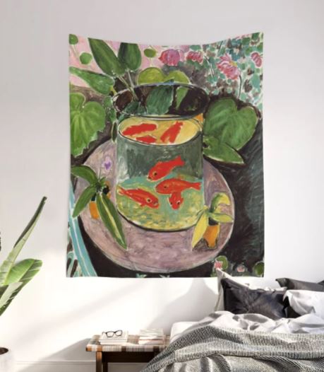 A vibrant piece from Blue Dog Posters' collection, the "Henri Matisse Goldfish Art Textile Small," adorns the wall, showcasing a bowl with several goldfish and lush green plants. Below, a portion of the bedroom's cozy atmosphere is revealed, featuring a bed with pillows alongside a wooden side table stacked with books. The rich colors infuse the room with an inviting ambiance reminiscent of colorful wall tapestries.