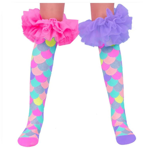 A close-up of a child's legs wearing Blue Dog Posters' Madmia Mermaid Frills Socks, featuring an overlapping scale pattern in shades of blue, pink, green, purple, and yellow. The child is also sporting bright pink and purple frilly tutu-like ruffled leg accessories at the thighs, adding a touch of glittery magic to the outfit.