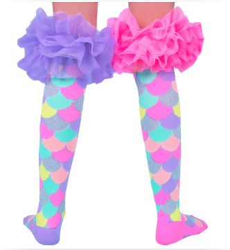 A child is wearing Madmia Mermaid Frills Socks by Blue Dog Posters, showcasing their colorful and glittering mermaid scale-patterned design with vibrant hues of pink, blue, green, and yellow. Over the socks are ruffled purple and pink shorts adorned with fluffy frills—a playful ensemble that brings these magical socks to life. The shot focuses on the legs and the lower part of the shorts.