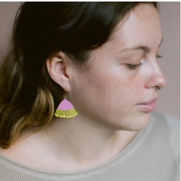 A woman with wavy hair looks downward, wearing Blue Dog Posters' Mini Gum Blossom Pink & Yellow Earrings—featuring a pink semicircle and yellow fringe—with a light top against a neutral background.