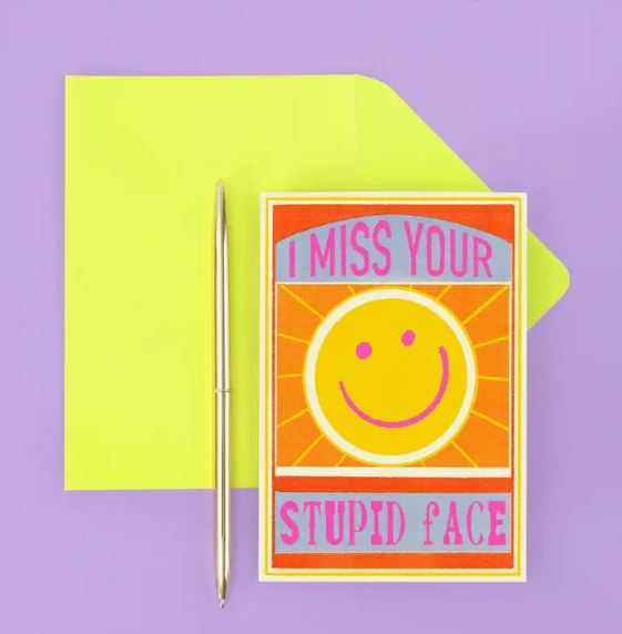 The Miss Your Stupid Face Greeting Card by Blue Dog Posters features a yellow smiley on an orange backdrop with "I Miss Your Stupid Face" in red and blue. It's paired with a yellow envelope and silver pen on a purple surface.