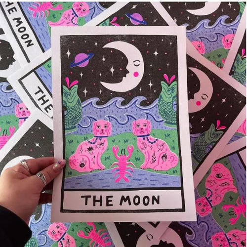 A hand holds "The Moon A4 Risograph - Amy Hastings" from Blue Dog Posters, showcasing a tarot card adorned with a crescent moon with a face, under a starry night sky. The card depicts mountains and a stream with a crab in the foreground, flanked by two pink creatures with human faces emerging from the water amidst green plants.