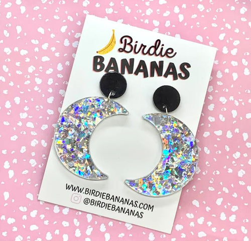 A pair of Moon Acrylic Earrings from Blue Dog Posters are displayed on a pink background with white speckles. Handmade in Australia, the earrings are mounted on a card featuring the brand's name and website, www.bluedogposters.com.