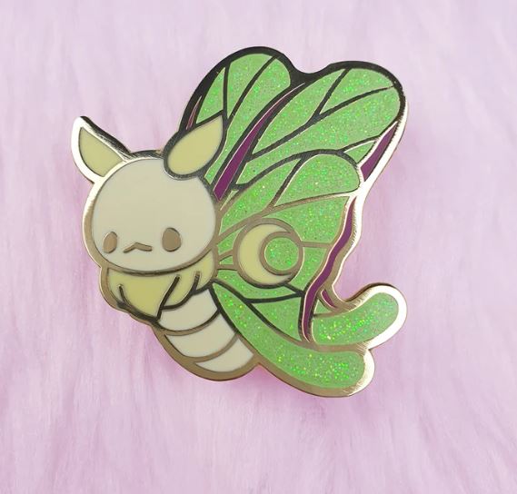 The Luna Moth Pin Pinstachiio by Blue Dog Posters showcases an adorable cartoon creature with yellow ears and wings that resemble shimmering green leaves. The small, round body and expressionless face of the moth are set against a soft pink fabric backdrop. Made from hard enamel, this pin features an enchanting original illustration.