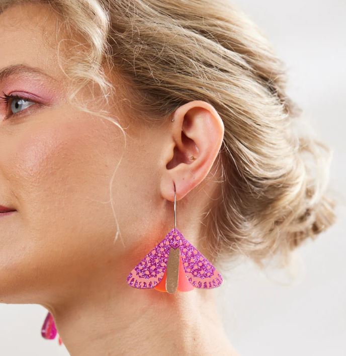 A close-up of a person with ear-length, tousled blonde hair wearing the vibrant Moth Earrings - Magenta Neon by Blue Dog Posters. The asymmetrical earring, which is orange and triangular with a textured glittery pattern, stands out prominently as the person looks slightly to the side.