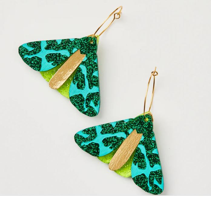 The Moth Earrings - Forest Green from Blue Dog Posters are beautifully showcased, with wings in forest green and teal adorned with shimmering leopard spots. Gold accents embellish their body, adding to their allure. Crafted in Australia, these captivating earrings elegantly dangle from delicate gold-colored hooks.