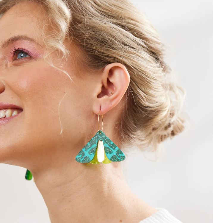 A woman with blonde hair smiles while wearing the Moth Earrings - Forest Green, showcasing vibrant shades of blue and forest green with a metallic finish. These earrings, crafted by Blue Dog Posters in Australia, beautifully complement her pink eyeshadow and white top against a softly blurred background.