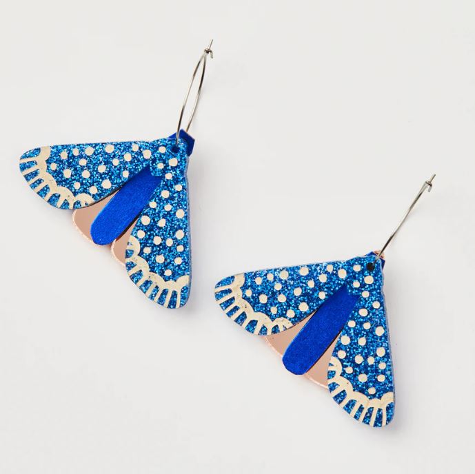 The Moth Earrings - Indigo by Blue Dog Posters are a pair of dangly earrings intricately crafted to resemble delicate moth wings, adorned with mesmerizing blue and beige patterns and decorated with sparkling accents, set against a plain white background. Created by Martha Jean in Australia, these earrings embody a unique blend of nature-inspired elegance.