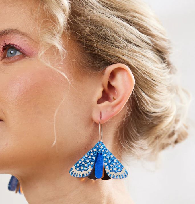 Close-up of a person with light-colored hair styled in a bun, wearing a large blue and black butterfly-shaped earring called the Moth Earrings - Indigo by Blue Dog Posters with intricate detailing. The person has pink eyeshadow and a small earring in the upper part of the ear, evoking the unique style seen in Australia.