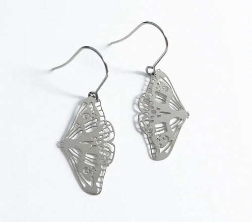 A pair of delicate, metallic butterfly-shaped earrings with intricate cut-out designs. Crafted from recycled plated stainless steel, the Denz Mini Earrings Moth Silver by Blue Dog Posters feature surgical steel hooks for a comfortable fit. The plain white background fully highlights the earrings' exquisite details.