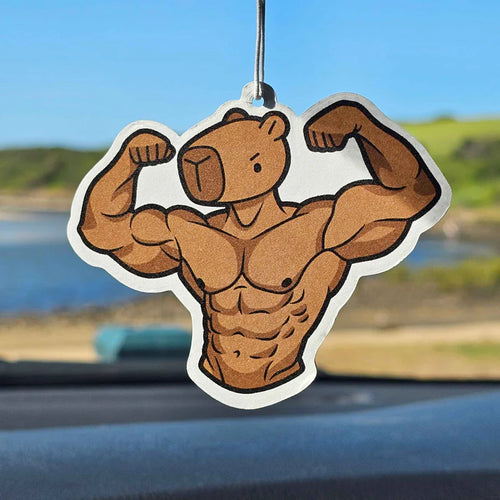 A Muscle Capybara Orange Air Freshener by Blue Dog Posters hangs from a car mirror, with a scenic landscape and blue sky visible through the windshield in the background.