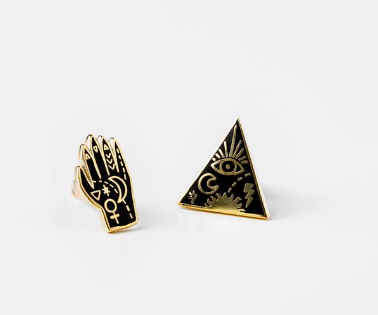 The Mystic Power Witchy Earrings by Blue Dog Posters feature a pair of black and gold enamel pins. One earring is shaped like a triangle adorned with symbols including an eye, sun, and celestial imagery, while the other takes the shape of a hand with matching intricate symbols such as a crescent moon, stars, and a Venus symbol, reminiscent of finely crafted cloisonné studs.