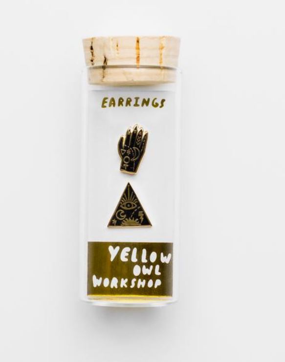 A pair of Mystic Power Witchy Earrings is displayed in a transparent container with a cork lid. One earring is a black hand with gold accents, and the other is a black triangle with an eye and gold accents. The label on the container reads "Blue Dog Posters." These cloisonné studs come with hypoallergenic posts for added comfort.