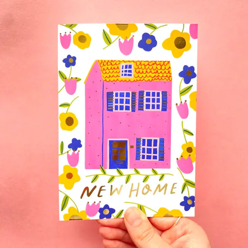 A hand holds the Blue Dog Posters New Home Pink House Greeting Card, featuring a pink house with blue shutters, yellow and purple flowers on a pink background. The card displays "NEW HOME" in gold letters at the bottom.