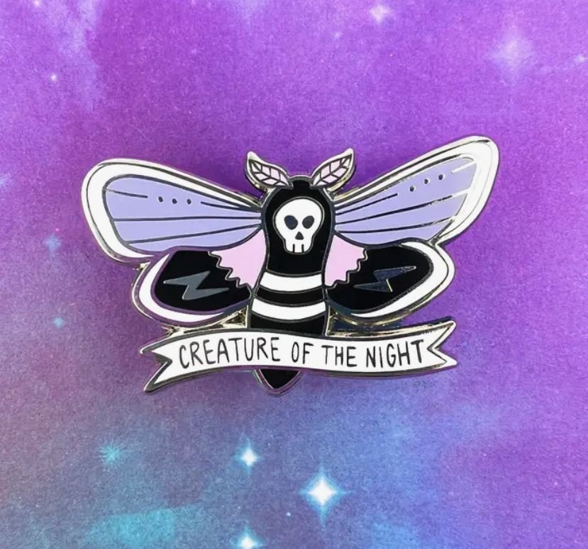Creature of the Night Enamel Pin by Blue Dog Posters, featuring a die struck metal design of a moth with a skull on its head and wings in shades of purple and black. The pin includes a banner reading "CREATURE OF THE NIGHT" against a background gradient of purple and blue, adorned with small star-like dots.