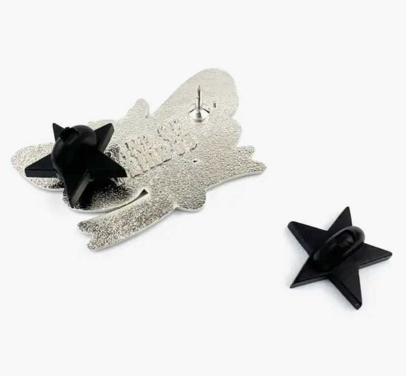 A metallic "Creature of the Night Enamel Pin" from Blue Dog Posters lies face down, revealing a single pin with a black rubber star-shaped backing clutched in place. Another identical enamel pin from the collection lies separately beside it on the pristine white surface.