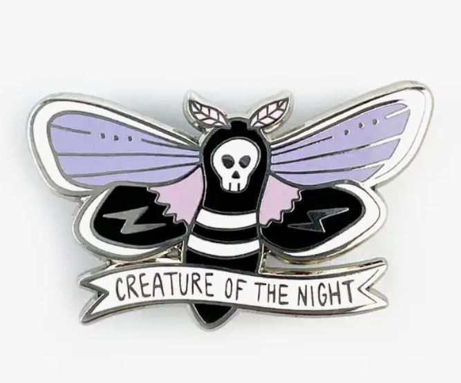 The Creature of the Night Enamel Pin by Blue Dog Posters features a stylized moth with a skull on its body, die struck in metal. This stunning piece showcases purple and black wings, intricate white detailing, and is set against a silver background. Below the moth, a white banner reads "CREATURE OF THE NIGHT.