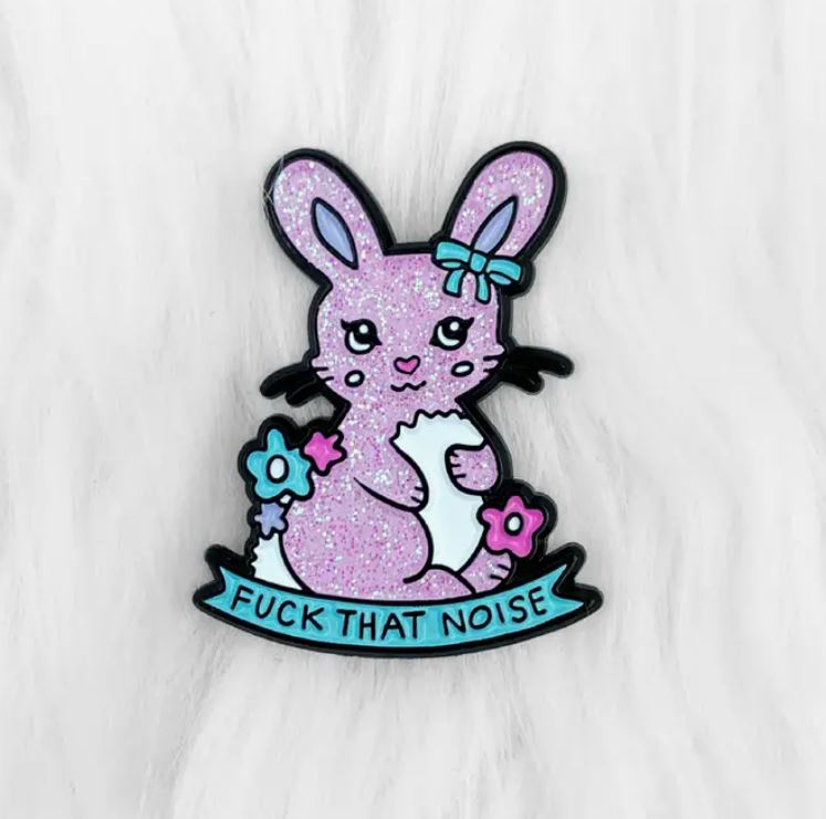 Presenting the Noise Rabbit Enamel Pin from Blue Dog Posters: a hard enamel pin showcasing an adorable pastel-colored bunny holding a flower and surrounded by floral accents. The bunny features a bow on its head and sparkling purple fur. Beneath the bunny, there's a ribbon with the text "FUCK THAT NOISE." The entire scene is set against a fluffy white background, designed to perfection by Blue Dog Posters.
