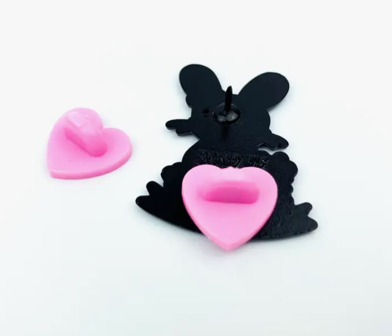 The Noise Rabbit Enamel Pin from Blue Dog Posters showcases a hard enamel pin with a silhouette of a black bunny. It features a single pin post and comes with a pink heart-shaped rubber clasp, along with an additional pink heart-shaped rubber clasp, all set against a white background.