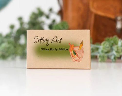 A small beige box with "Getting Lost Cards - Office Party Edition" by Blue Dog Posters written on it. There is a drawing of a drink with a garnish on the right side. The background is slightly blurred, featuring some greenery and a brown leather bag—perfect for team bonding at any office party.