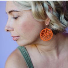 A person with short, wavy blonde hair and a sleeveless green top is shown in profile against a purple background, wearing Denz Citrus Slice Earrings in Orange by Blue Dog Posters.