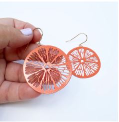 A hand displays the Denz Citrus Slice Earrings in Orange by Blue Dog Posters, featuring intricate abstract floral cut-out patterns. The backdrop is a plain, white surface.